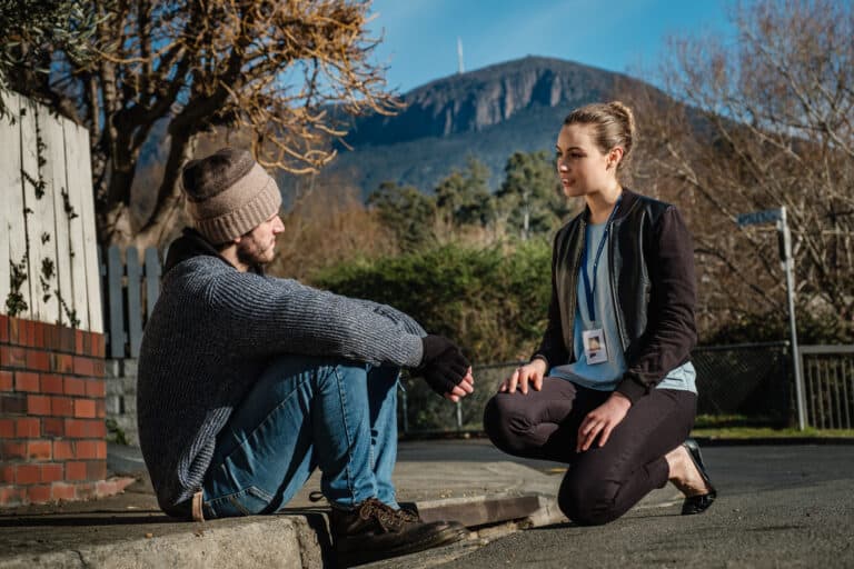 A man in a beanie sits on a footpath and a support worker kneels beside him